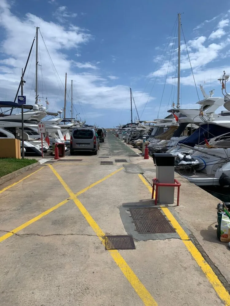 A marina walkway lined with parked yachts on both sides under a partly cloudy sky.