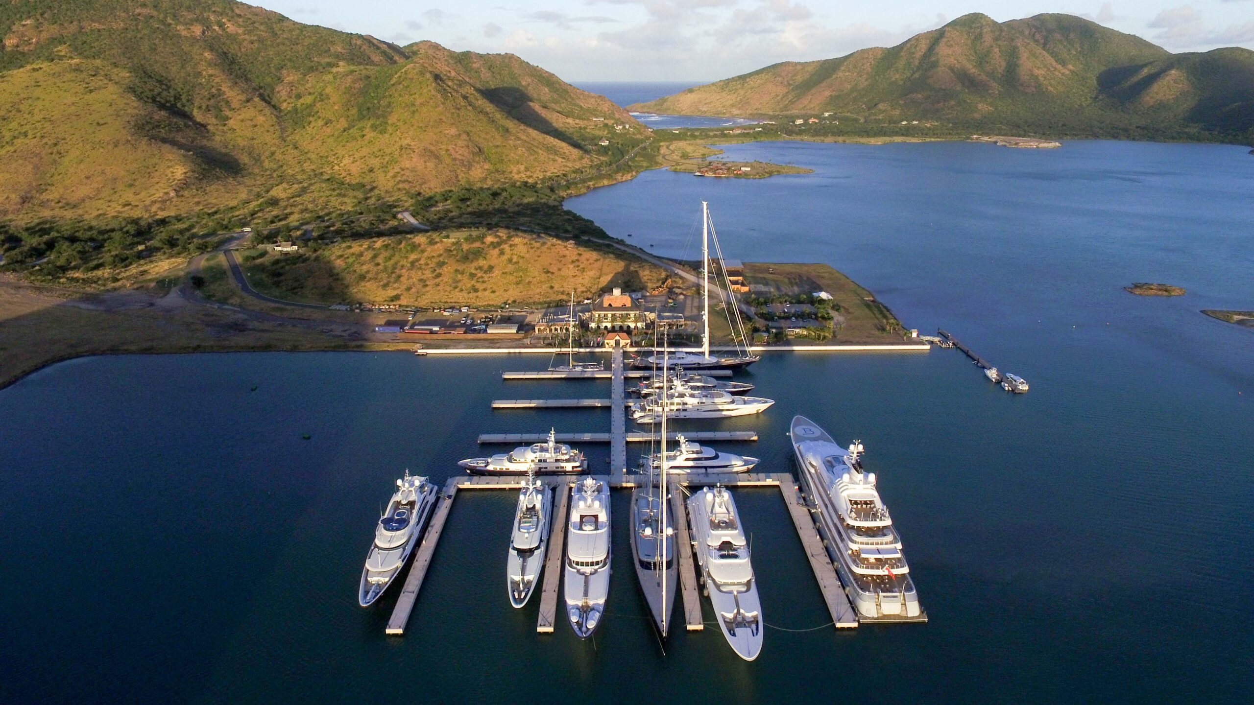 Aerial view of a marina with luxury yachts docked, surrounded by green mountains and calm blue water.
