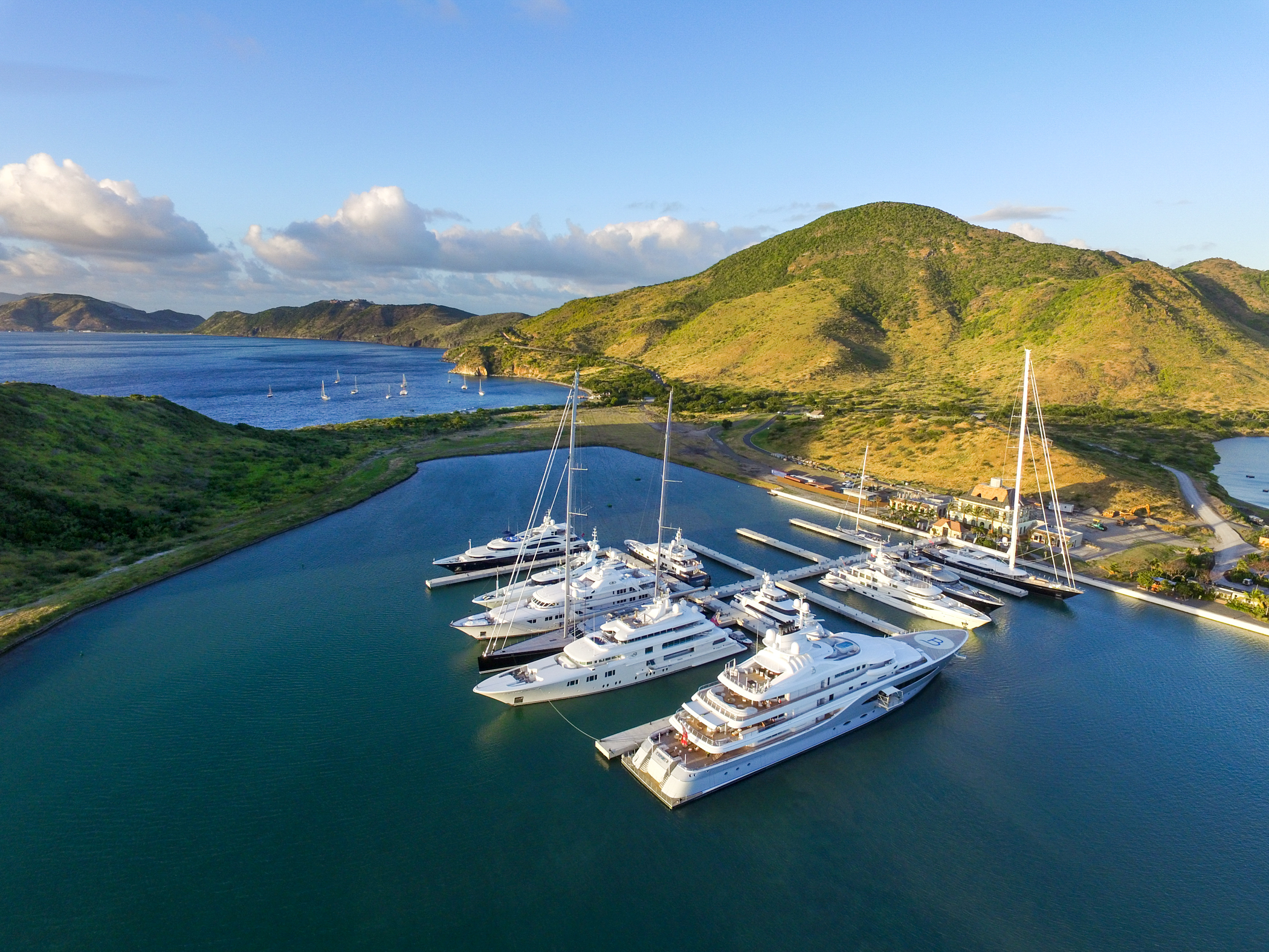 Luxury yachts docked at a marina surrounded by green hills and a calm blue sea under a clear sky.