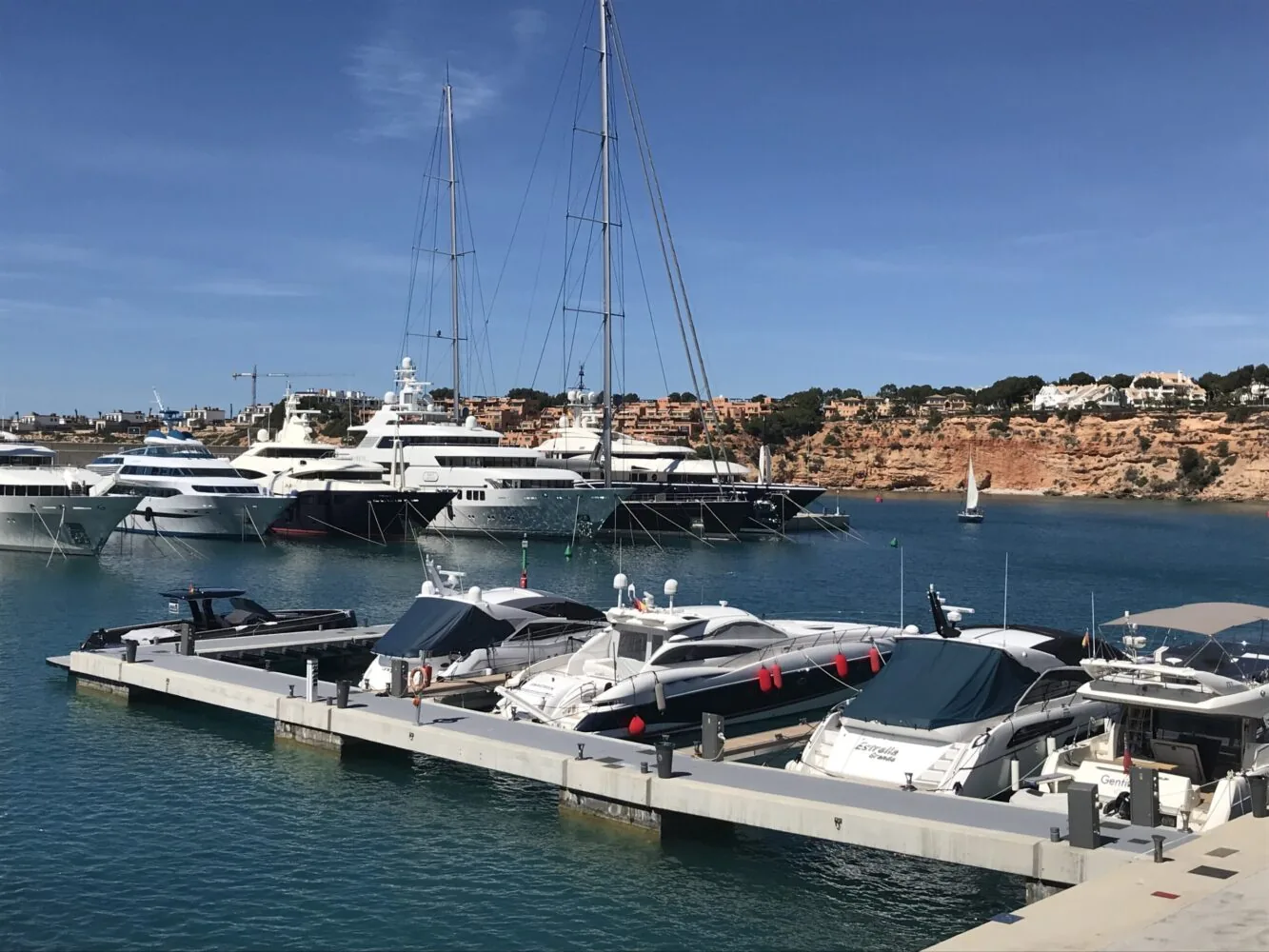 Luxury yachts docked at a marina with cliffs and houses in the background under a clear blue sky.