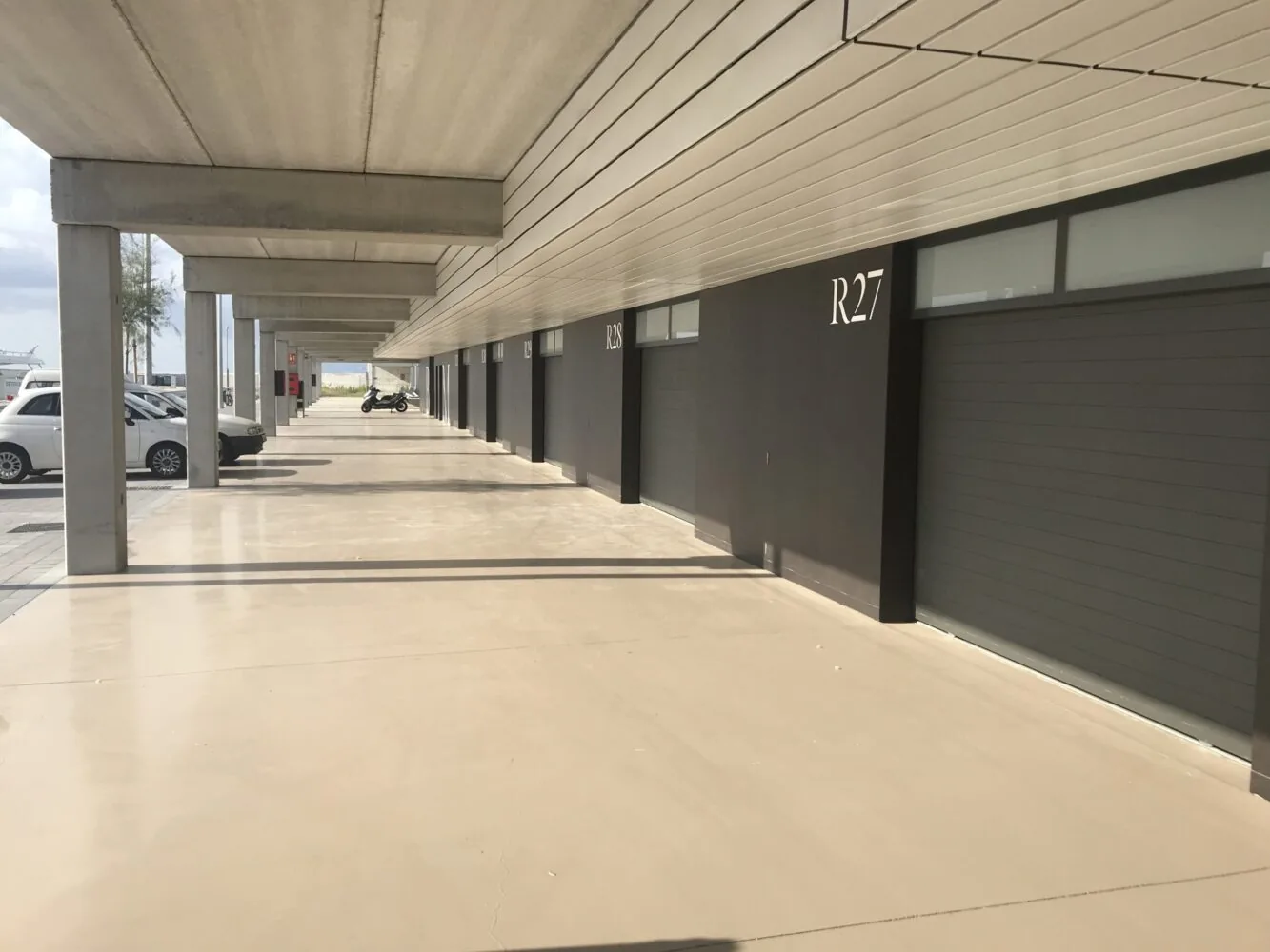 Covered walkway next to a row of closed garage units labeled "R27" with parked vehicles and a scooter in the distance.