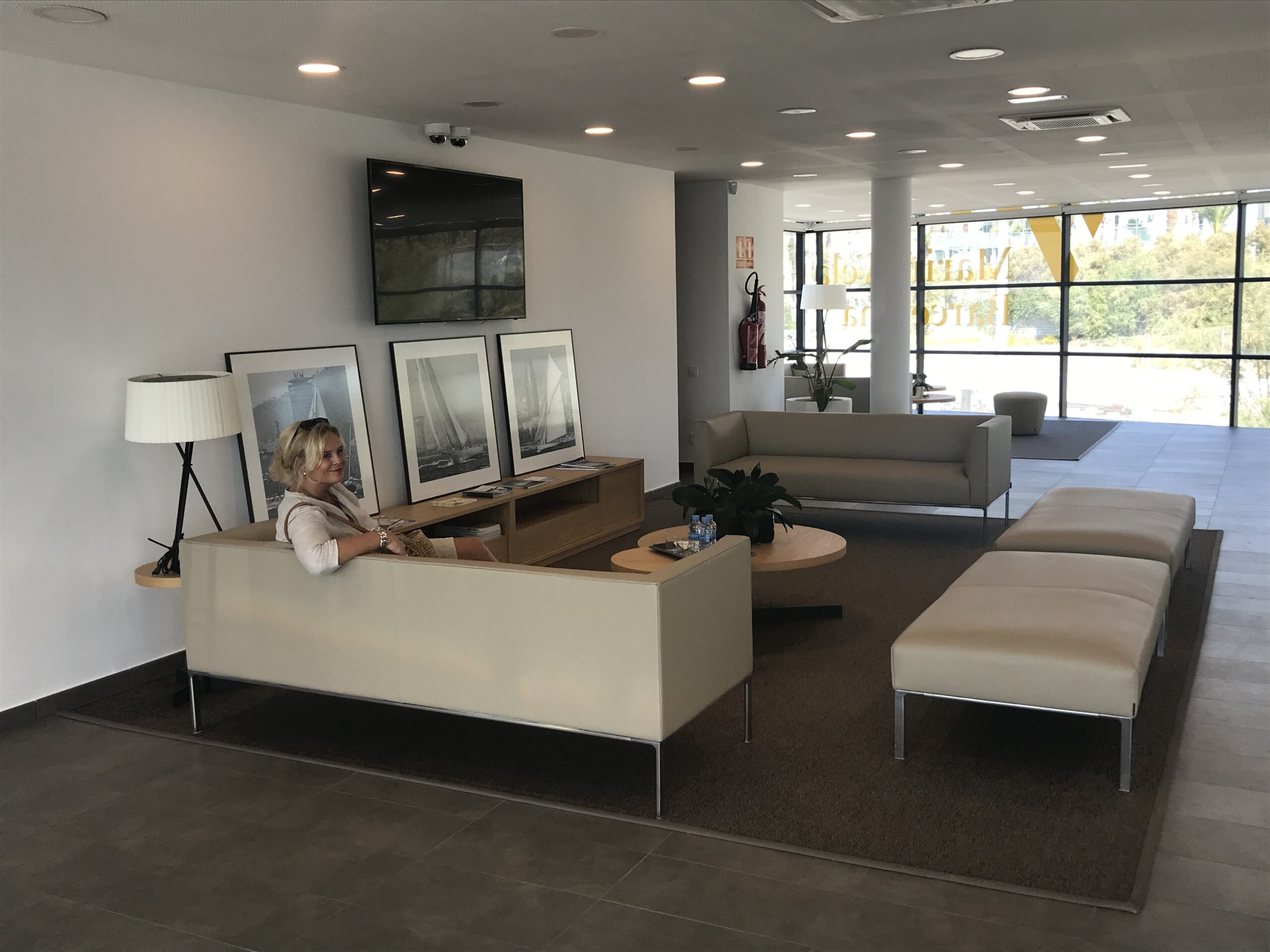 Modern waiting area with beige furniture, large windows, and a woman sitting on a sofa near framed nautical photographs.