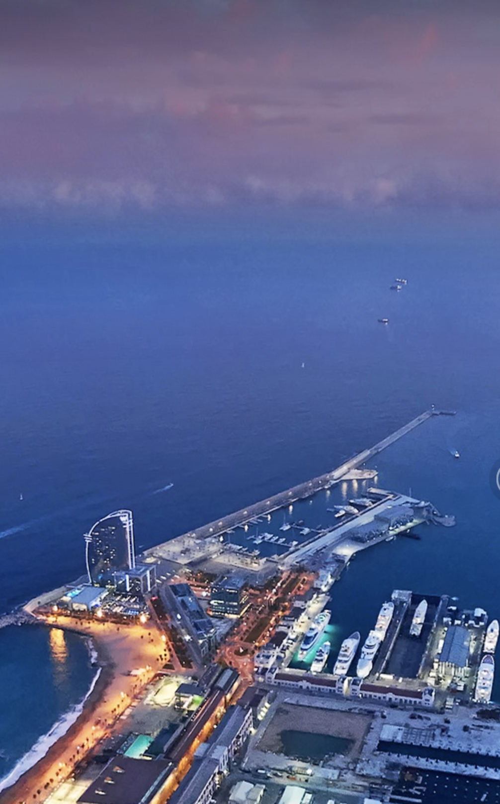 Aerial view of Barcelona's coastline at dusk, featuring the W Hotel, marina, and surrounding city lights.
