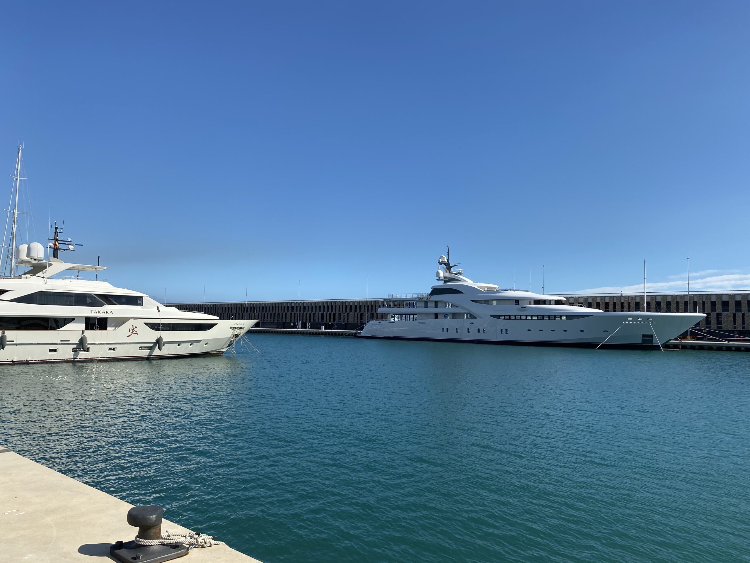 Two luxury yachts, including one named "TAKARA", docked in a calm marina under a clear blue sky.
