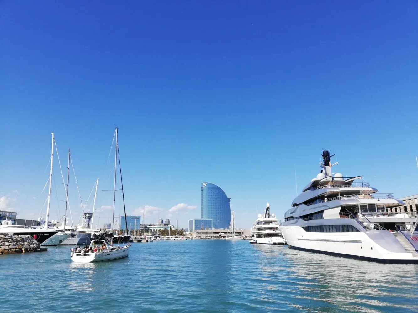Luxusjachten und Segelboote, die in einem Jachthafen angedockt sind, mit dem W Barcelona Hotel im Hintergrund unter einem klaren blauen Himmel.