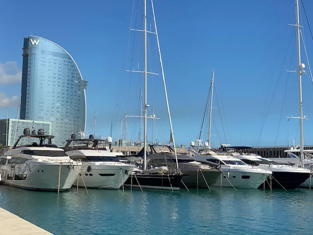 Luxury yachts docked in a marina with the W Barcelona hotel visible in the background under a clear blue sky.