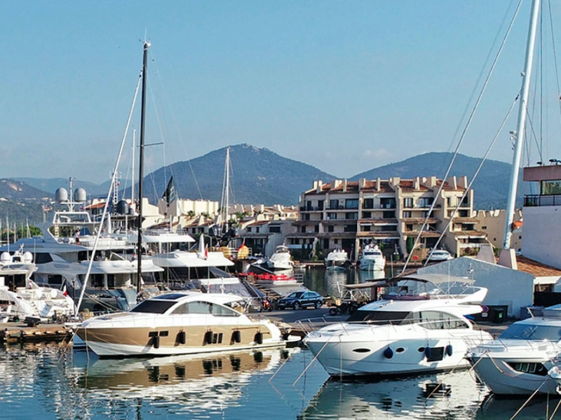 Luxury yachts docked at a marina in Marines de Cogolin, France, with residential buildings and hills in the background.