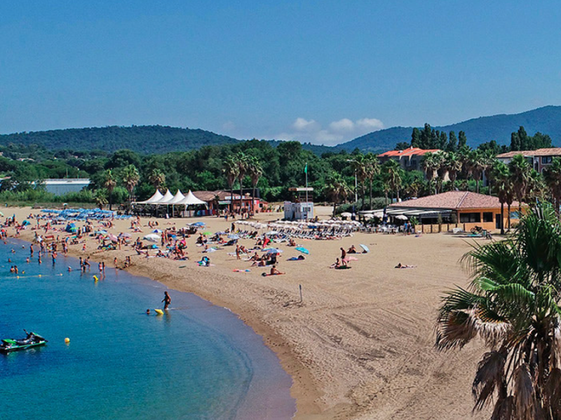 Busy beach scene at Marines de Cogolin in France with sunbathers, beach facilities, and surrounding hills.