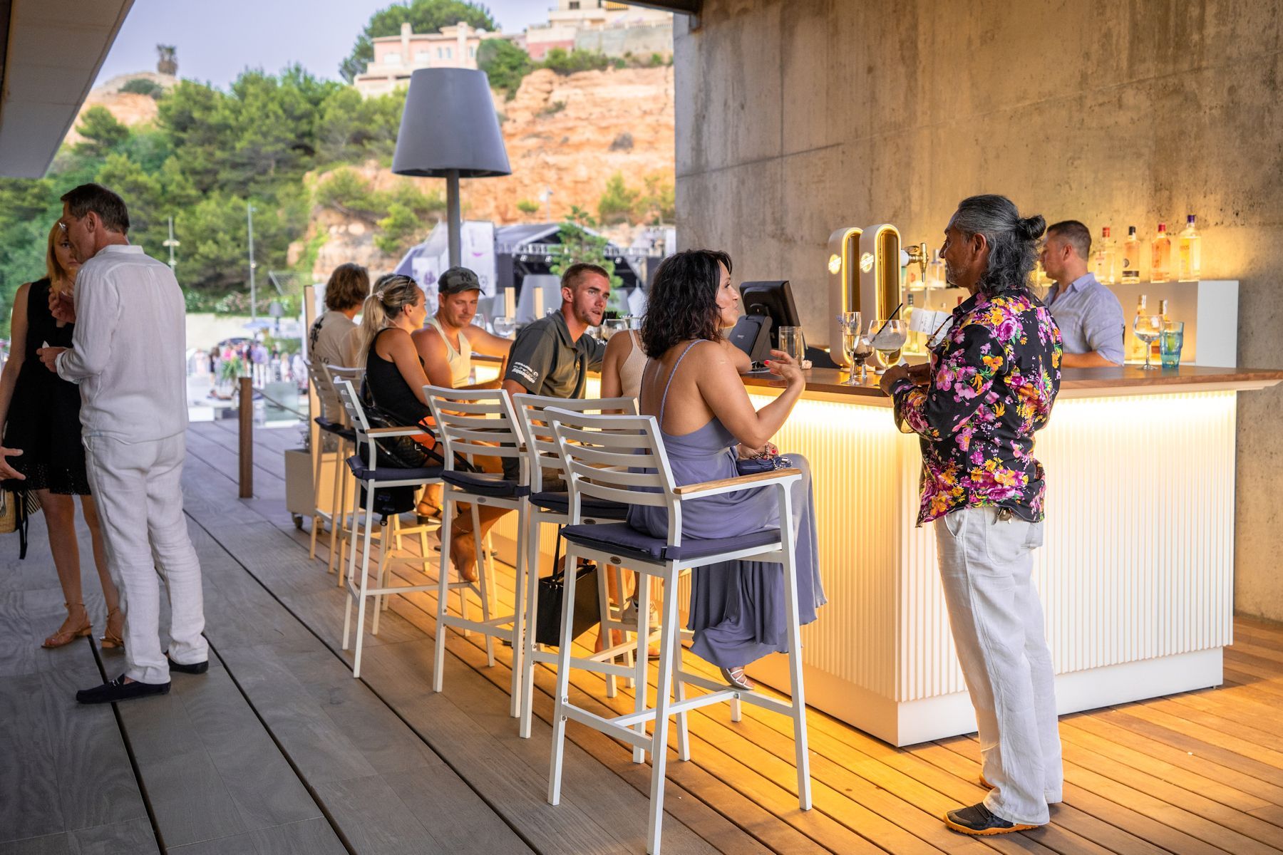 People socializing and enjoying drinks at an outdoor bar at PA Restaurants with a scenic hillside view in the background.