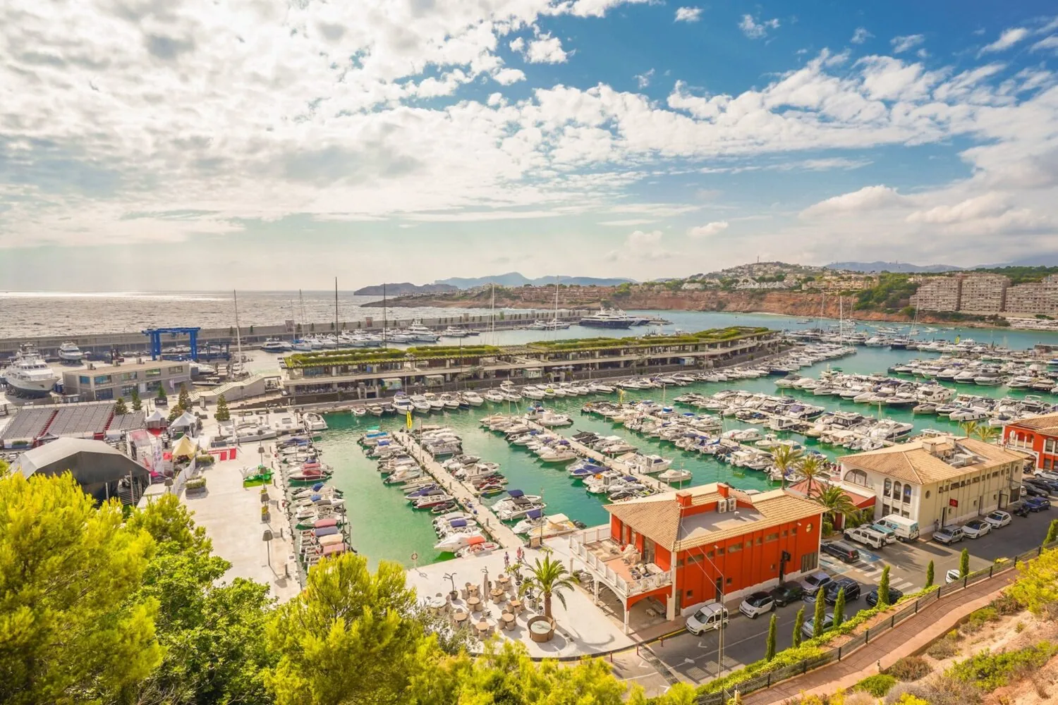 Aerial view of Port Adriano marina with numerous yachts, turquoise water, and surrounding buildings on a sunny day.