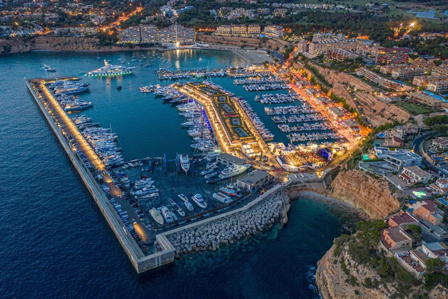 Aerial view of Port Adriano at dusk, showcasing a marina filled with yachts, illuminated waterfront buildings, and surrounding cliffs and residential areas.
