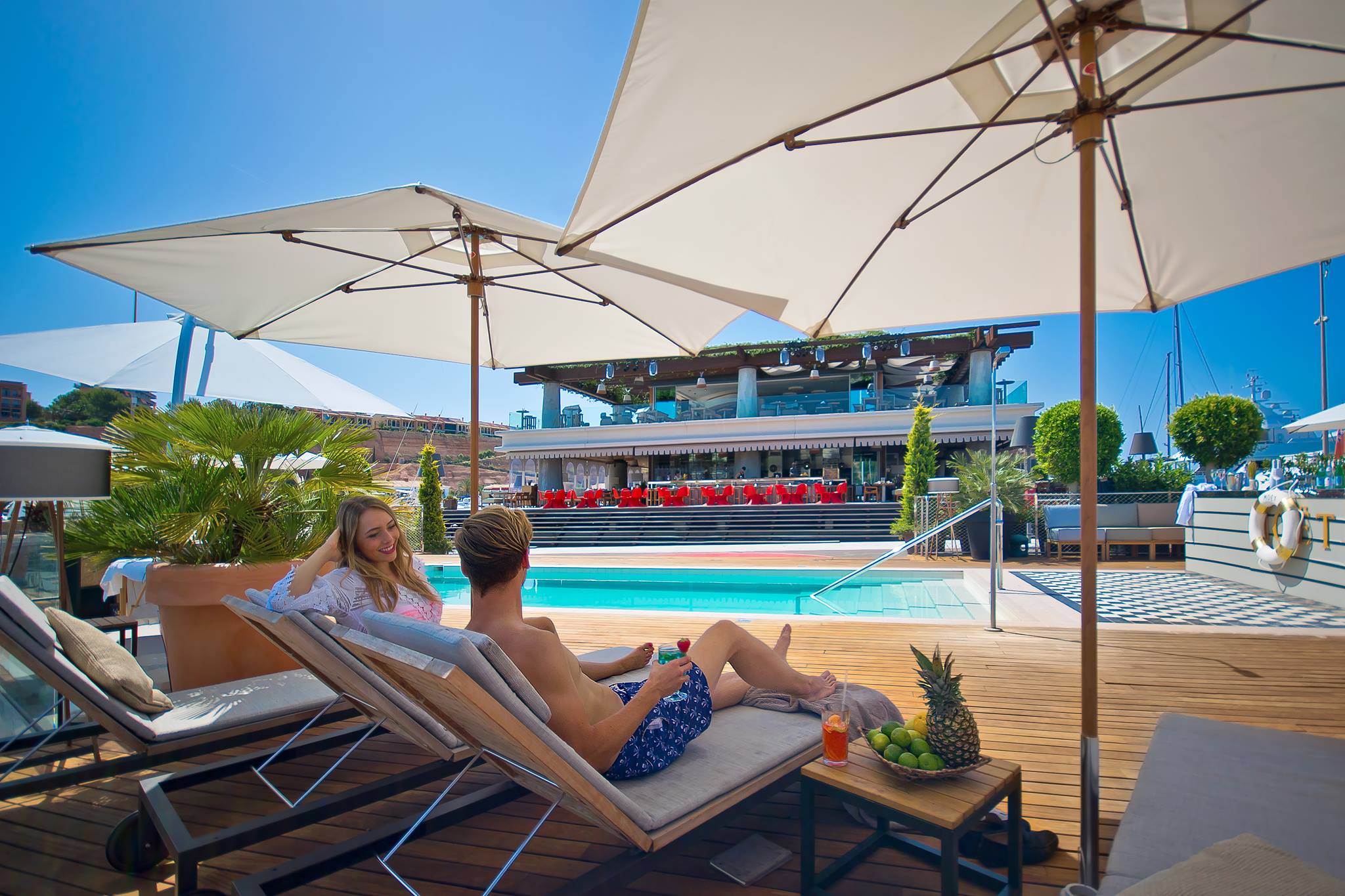 Couple relaxing on sun loungers under umbrellas by the luxurious PA pool with drinks and tropical fruit.