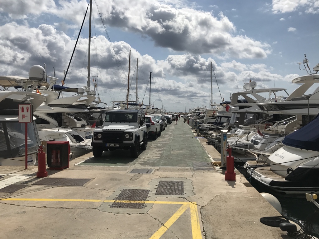 A narrow quay at Portals Quay 15-18M lined with luxury yachts and parked vehicles under a partly cloudy sky.