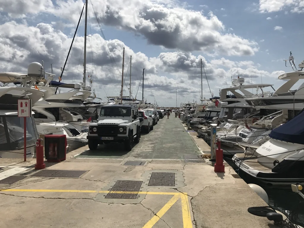 A narrow quay at Portals Quay 15-18M lined with luxury yachts and parked vehicles under a partly cloudy sky.