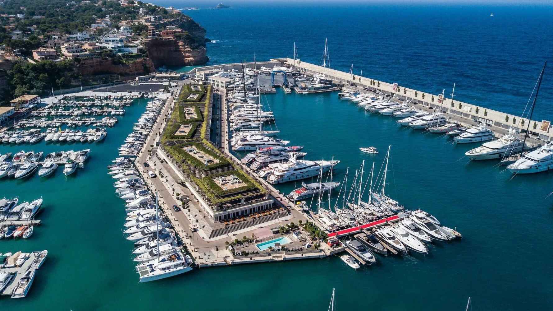Aerial view of Port Adriano marina in 2019, showcasing luxury yachts, waterfront buildings, and surrounding coastal landscape.