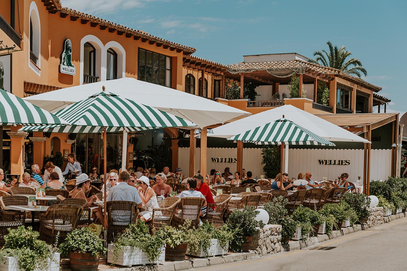 People dining outdoors at the Wellies restaurant terrace in Portals Nous under green and white striped umbrellas.