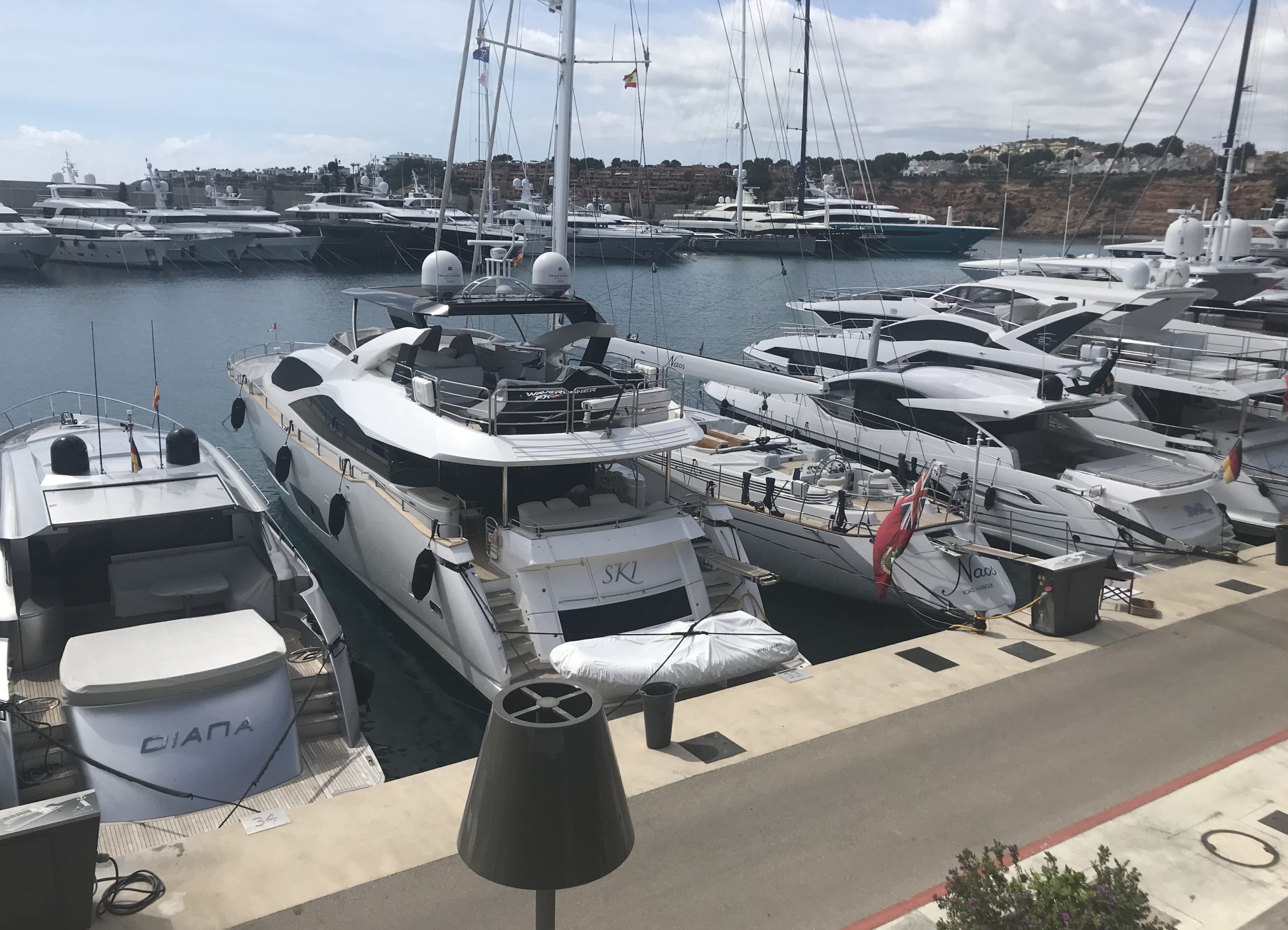 Luxury yachts docked in a marina, including the boats named "DIANA," "SKL," and "Naos," under a partly cloudy sky.