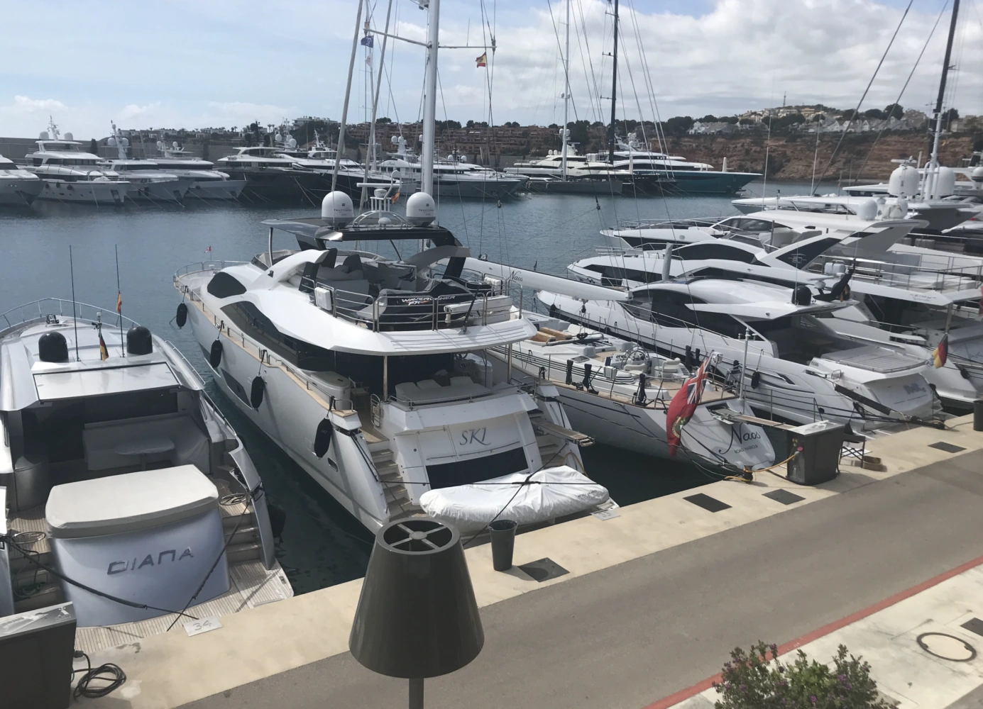 Luxury yachts docked in a marina, including the boats named "DIANA," "SKL," and "Naos," under a partly cloudy sky.