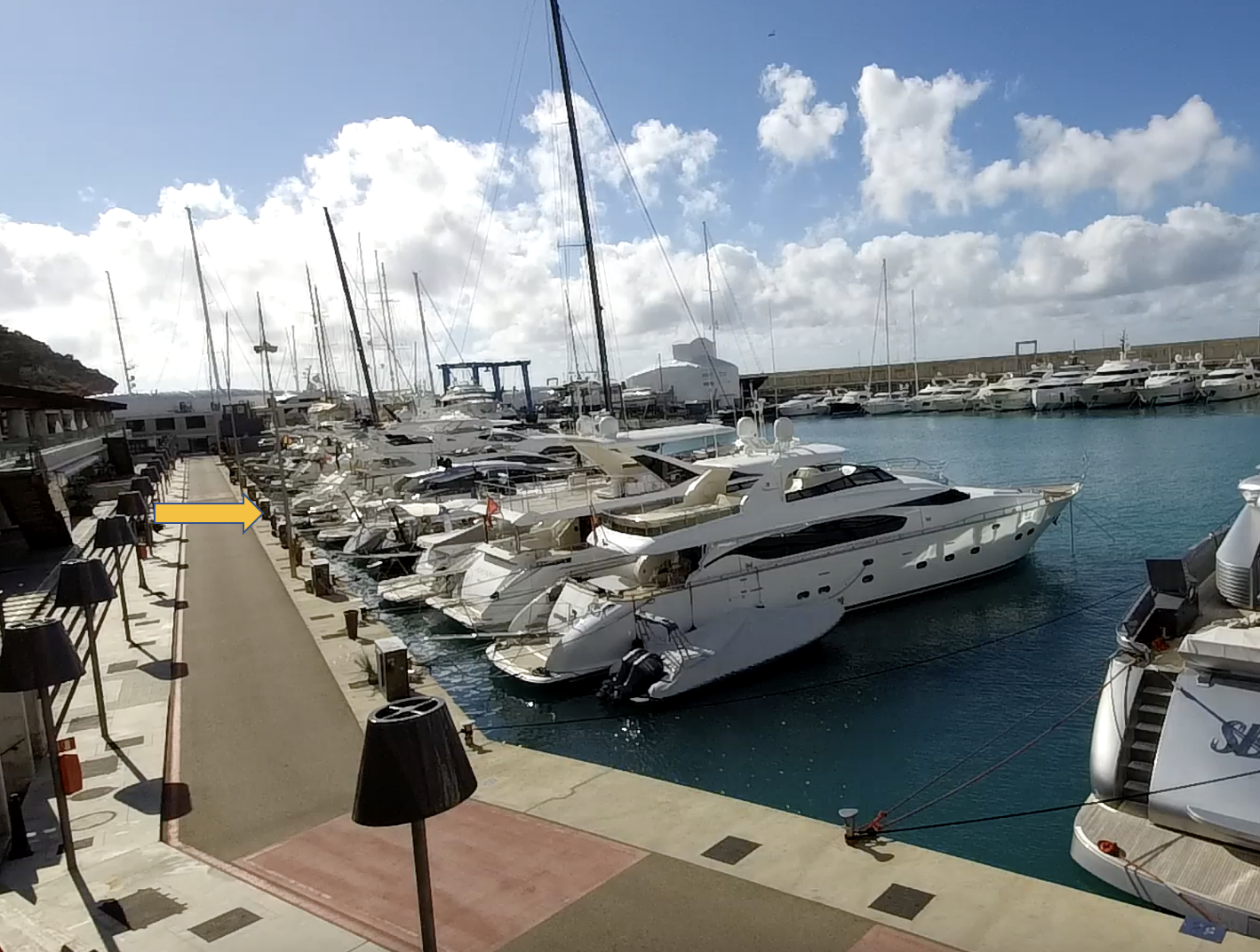 Yachts moored along a marina dock under a partly cloudy sky with a walkway running beside them.