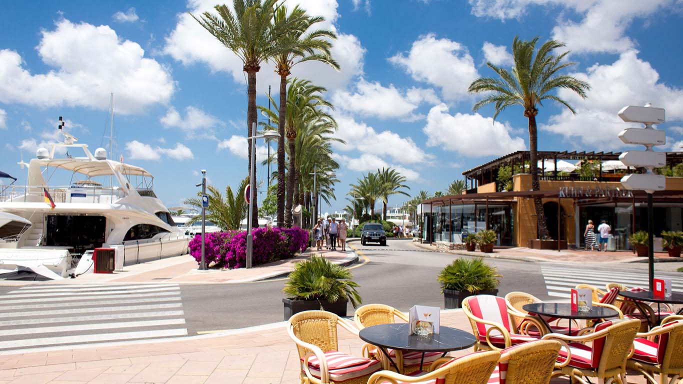 Outdoor café with red-striped chairs overlooking luxury yachts and palm-lined streets in Portal Nous.