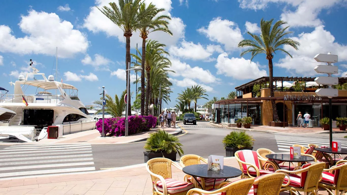 Outdoor café with red-striped chairs overlooking luxury yachts and palm-lined streets in Portal Nous.