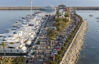 Eine lebhafte Yachthafenszene in Portals, in der auf der einen Seite Luxusyachten vor Anker liegen und Menschenmengen entlang einer von Palmen gesäumten Promenade zwischen geparkten Sportwagen spazieren.