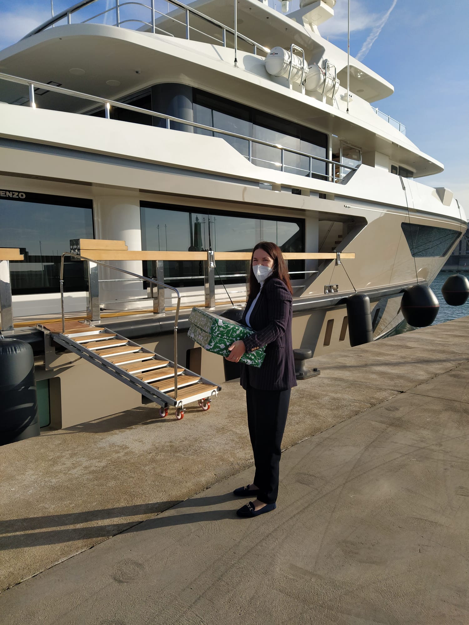 Woman wearing a face mask holding a wrapped gift box in front of a luxury yacht docked at a marina.