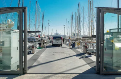 Ein Transporter fährt durch offene Glastore in einen Yachthafen ein, an dessen beiden Seiten unter einem strahlend blauen Himmel Yachten und Segelboote vor Anker liegen.