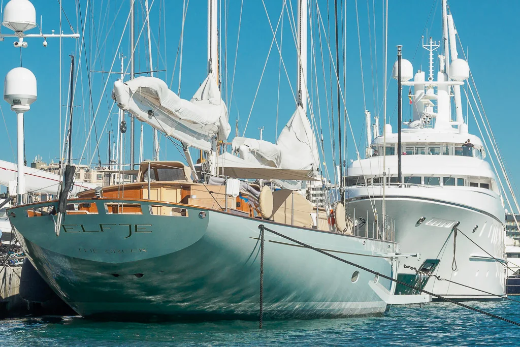 Two luxurious yachts, including the sailboat "Elfje," are moored side by side in a marina under clear blue skies.