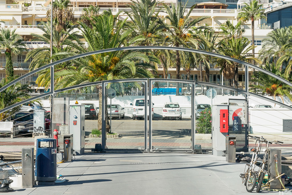 Entrance gate to a parking area with palm trees and a multi-story building in the background.
