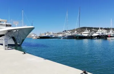 Luxury yachts moored in a sunny marina with clear blue water and a hilly backdrop.