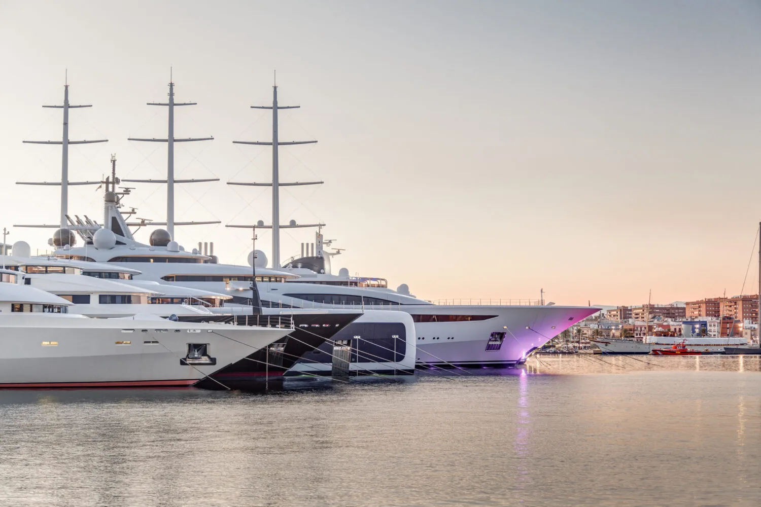 Luxury yachts docked in a marina at sunset with city buildings in the background.