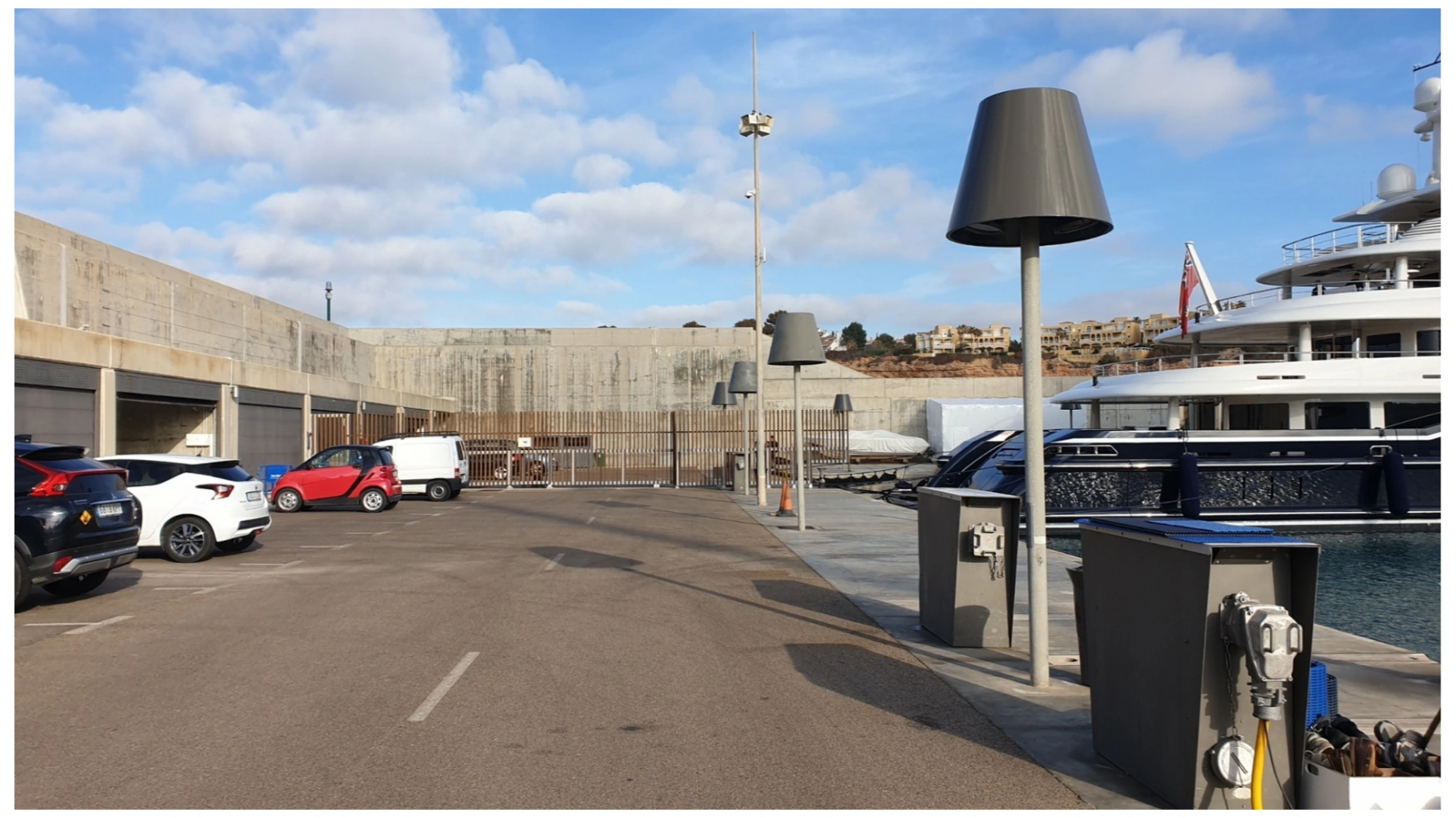 Parking area at Port Adriano Berth 79 with several cars and a large yacht docked alongside.