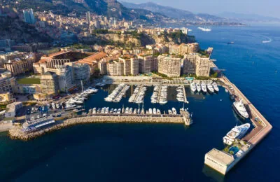 Aerial view of Port de Cap d’Ail with luxury yachts, waterfront buildings, and the mountainous coastline of Monaco in the background.