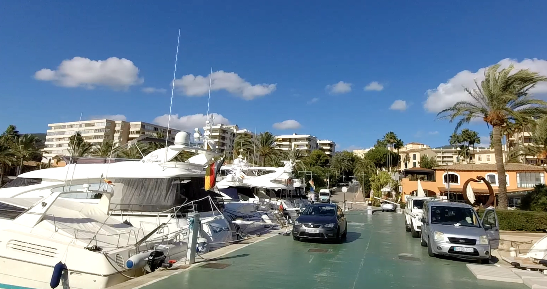 Luxury yachts docked at a sunny marina surrounded by palm trees, parked cars, and coastal apartment buildings.
