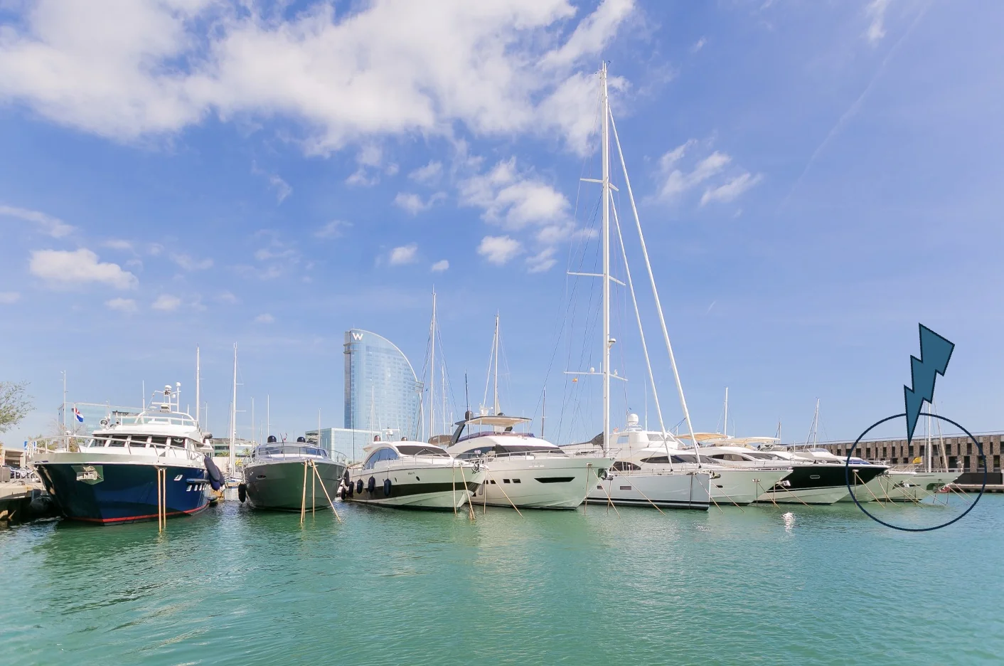 A variety of yachts docked at a marina with a modern glass building in the background under a clear blue sky.