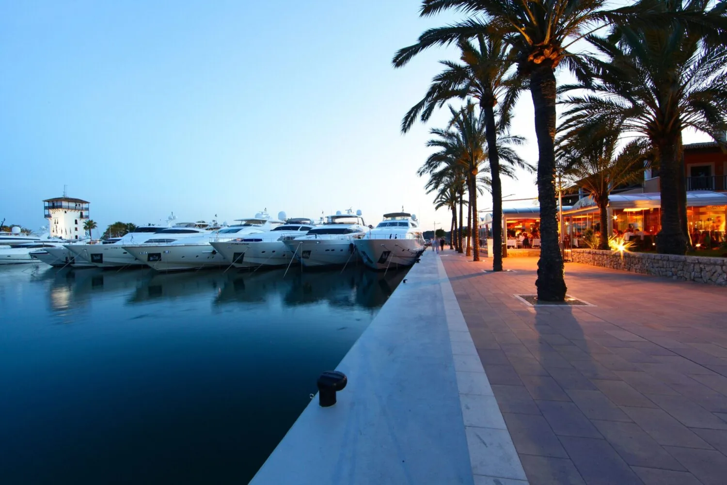 Bei Sonnenuntergang lagen Luxusyachten in einem Yachthafen neben einer von Palmen gesäumten Promenade mit Restaurants vor Anker.