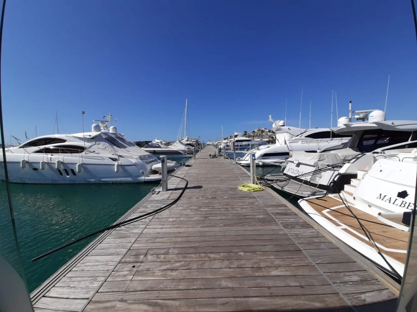 Wooden dock lined with luxury yachts on both sides under a clear blue sky.