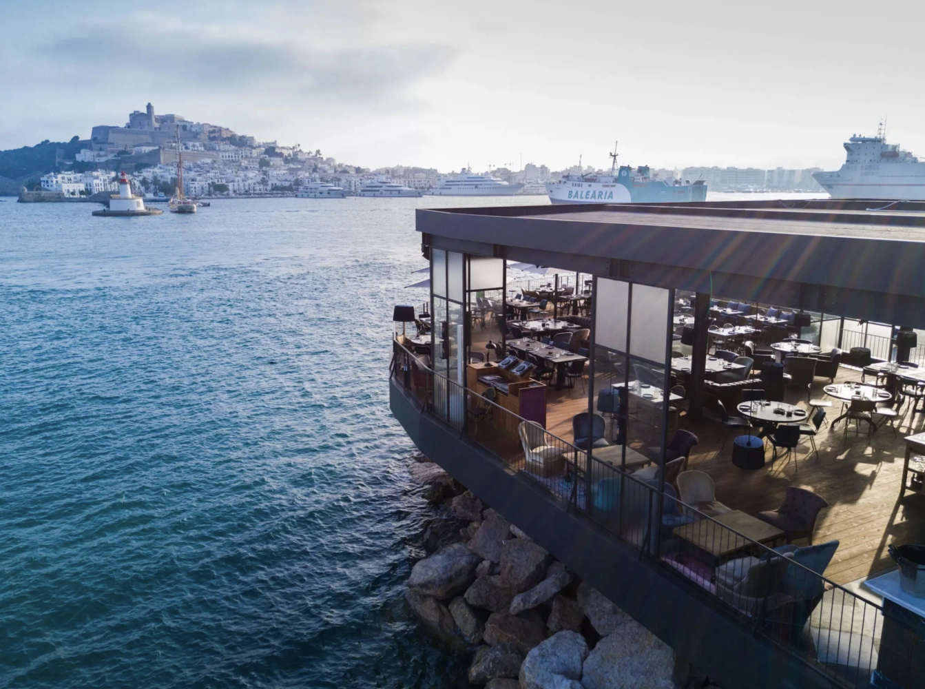 A restaurant with large windows overlooks the sea in Ibiza, with the island's historic architecture and ferries in the background.
