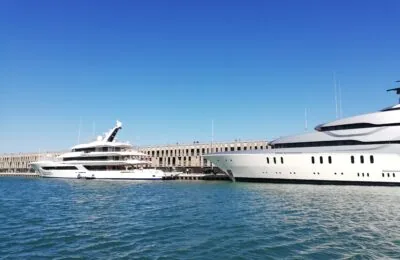 Two luxurious white yachts docked in a harbor with a large stone building in the background under a clear blue sky.