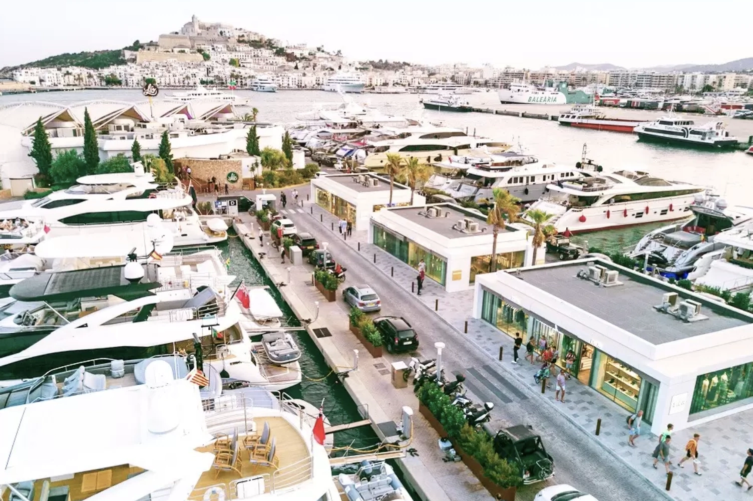 A vibrant marina in Ibiza with luxury yachts docked, surrounded by shops and palm trees, with a view of the town on the hill in the background.