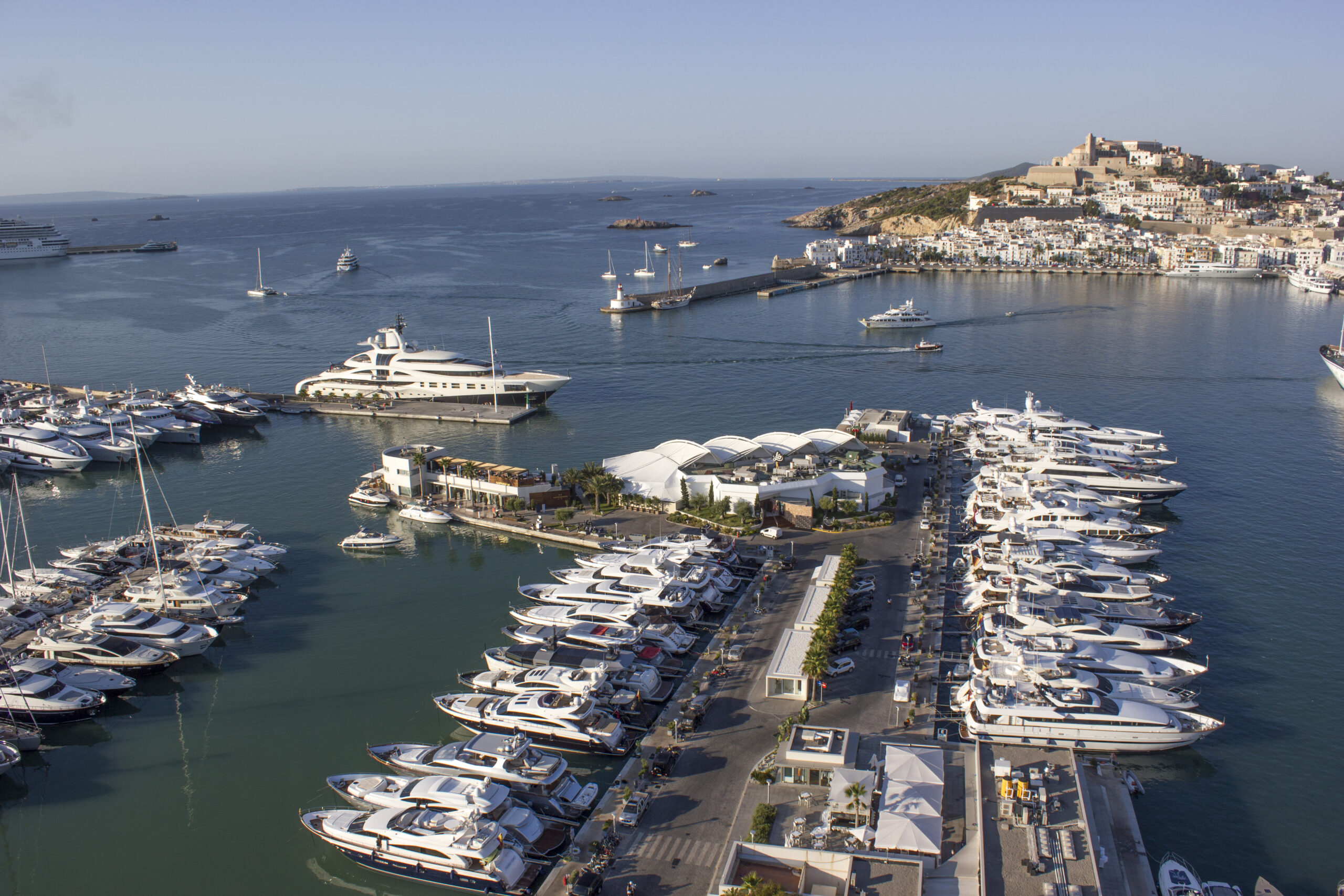 Aerial view of a luxury marina filled with yachts in Ibiza, with the old town and fortress visible in the background.