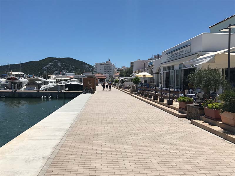 A sunny promenade in Santa Eulalia with yachts docked on the left and restaurants on the right.
