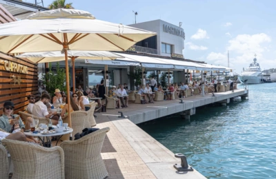 Die Außenterrasse des „Cappuccino Grand Café“ am Yachthafen, auf der sich an einem sonnigen Tag zahlreiche Gäste versammelt haben, um am Wasser einen Drink zu genießen.