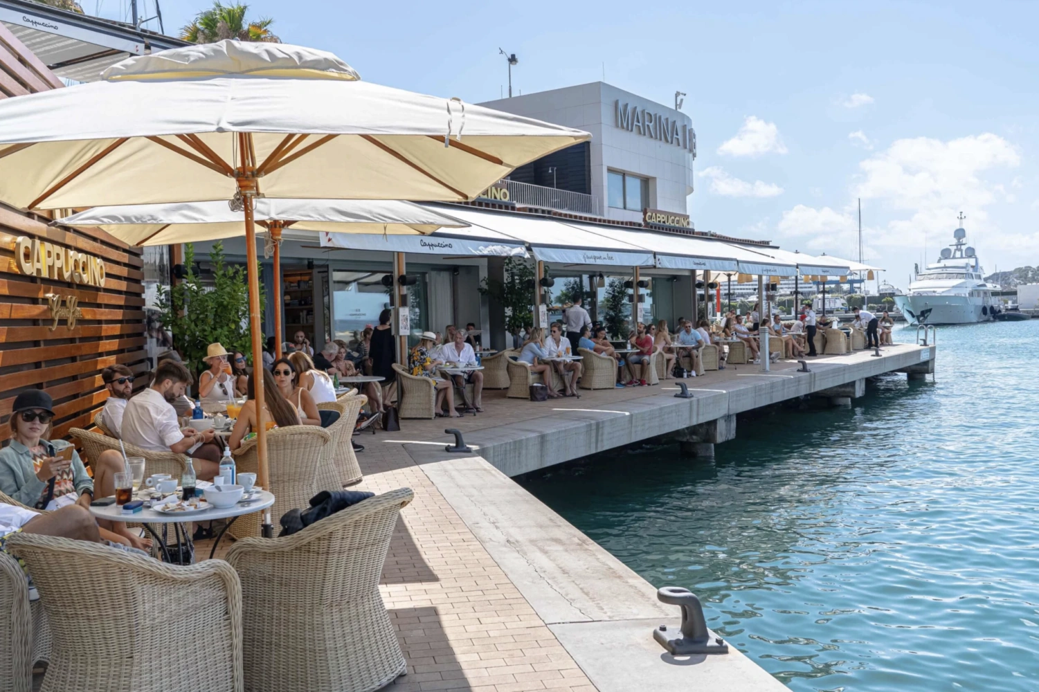 Die Außenterrasse des „Cappuccino Grand Café“ am Yachthafen, auf der sich an einem sonnigen Tag zahlreiche Gäste versammelt haben, um am Wasser einen Drink zu genießen.