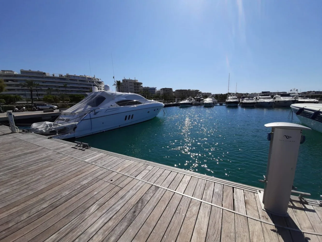 A white luxury yacht docked at a sunny marina with clear blue water and modern buildings in the background.