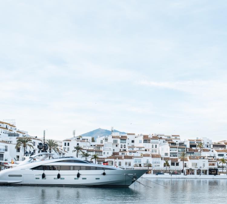 Modern yacht docked in a calm marina with white Mediterranean-style buildings and mountains in the background.