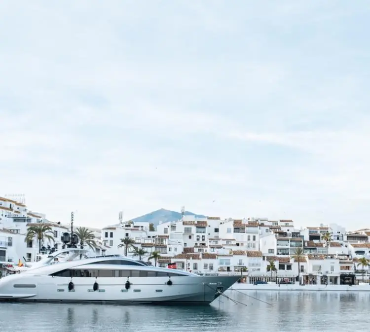 Modern yacht docked in a calm marina with white Mediterranean-style buildings and mountains in the background.