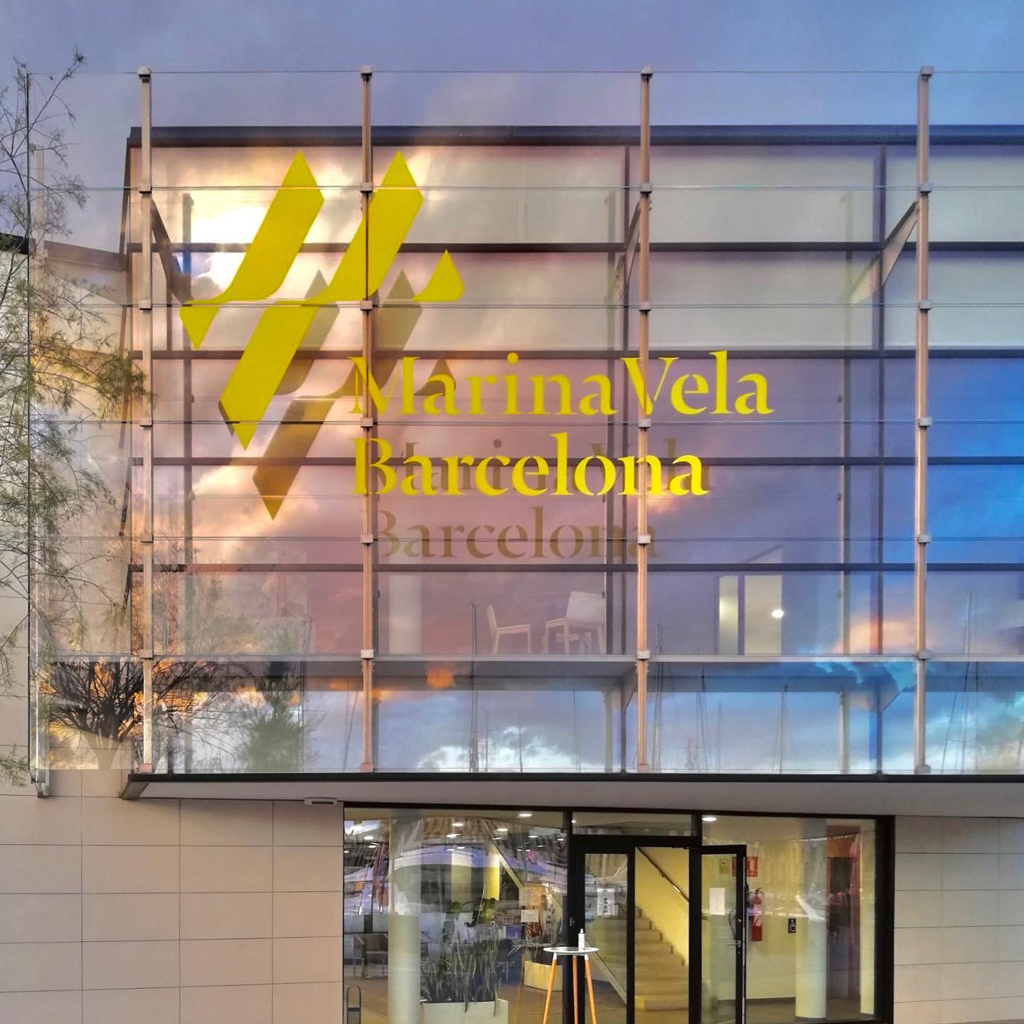 Glass building facade of Marina Vela Barcelona reflecting a colorful sunset sky.