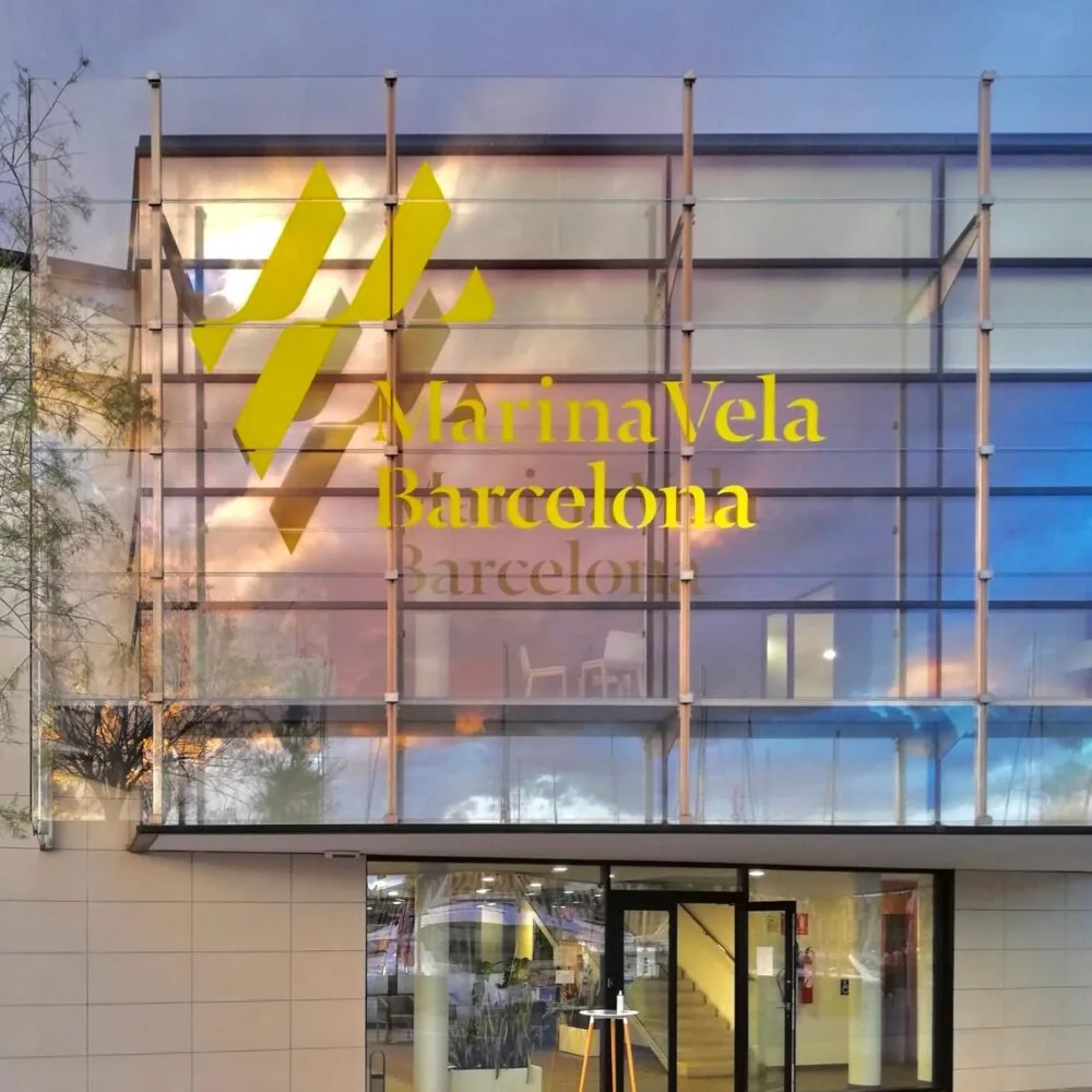 Glass building facade of Marina Vela Barcelona reflecting a colorful sunset sky.